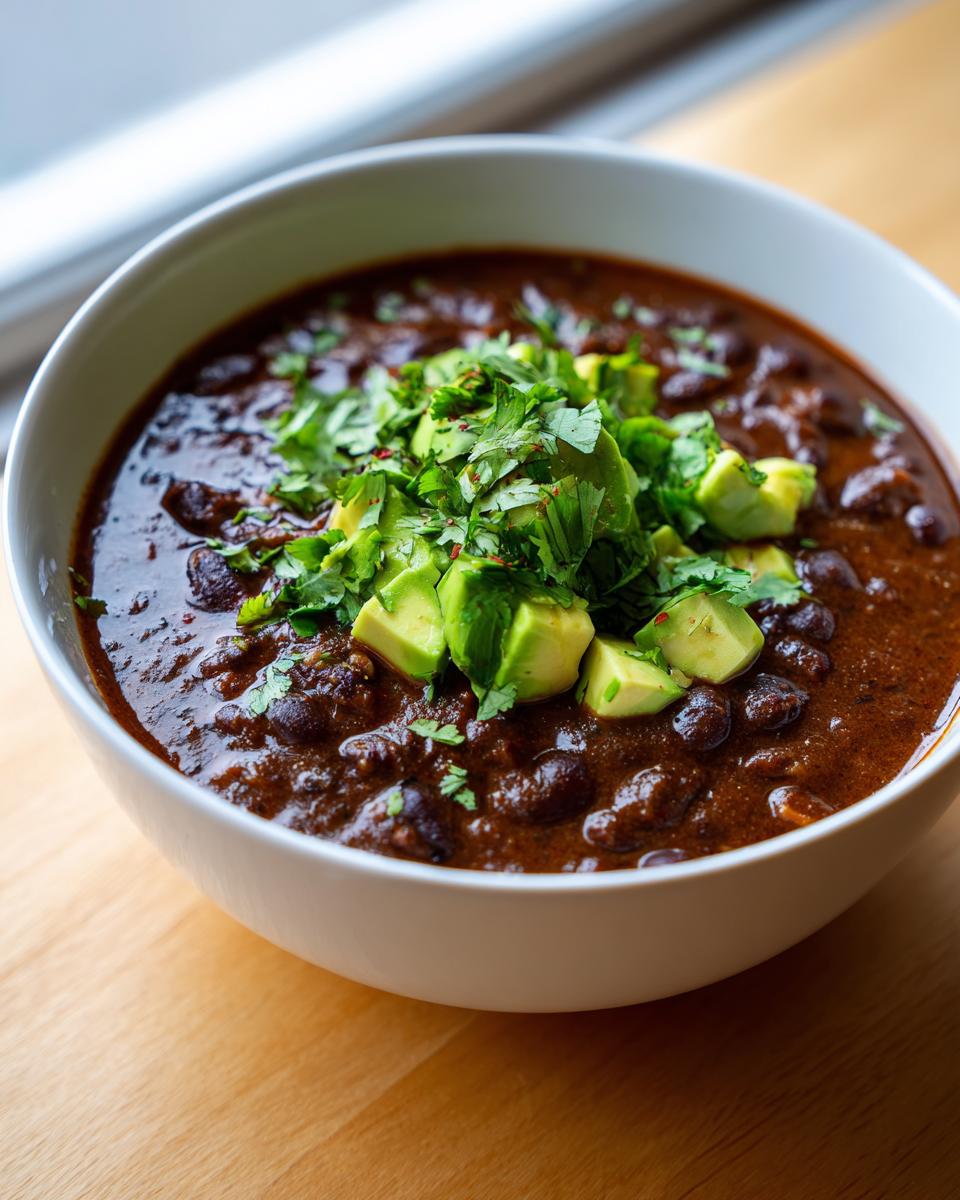 A close-up of a white bowl filled with rich, dark spicy black bean soup, garnished generously with diced avocado and fresh cilantro.