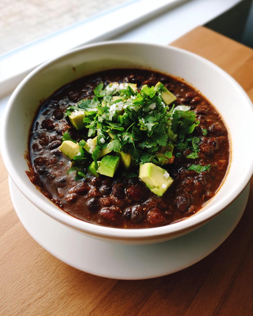A bowl of rich, dark spicy black bean soup garnished generously with diced avocado, fresh cilantro, and cotija cheese.