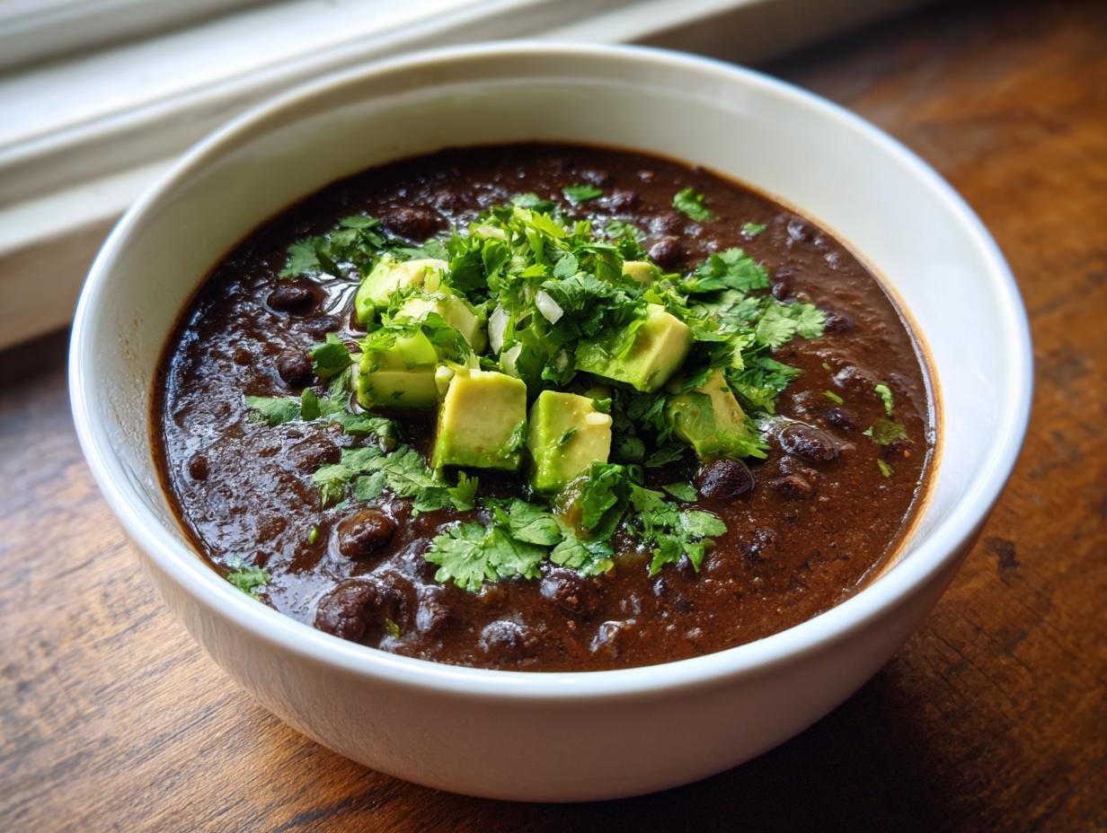 A close-up of a hearty bowl of spicy black bean soup, garnished generously with diced avocado and fresh cilantro.