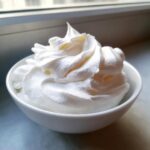 Close-up of stiff, bright white royal icing recipe mixture piped into soft peaks in a small white bowl.