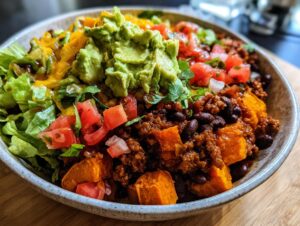 A close-up of a vibrant sweet potato taco bowl filled with seasoned ground meat, black beans, diced tomatoes, shredded lettuce, and guacamole.