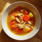 A close-up of a bowl of steaming chicken vegetable soup featuring shredded chicken, carrots, and potatoes in a savory broth.
