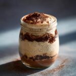 A layered tiramisu chia pudding in a glass jar, topped with cocoa powder.