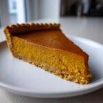 Close-up of a perfect slice of vegan pumpkin pie with a thick, vibrant orange filling and a crumbly crust.