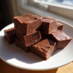 A stack of rich, dark chocolate squares of homemade fudge piled on a white plate.