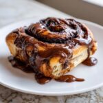 Close-up of a single, gooey chocolate cinnamon rolls smothered in rich, melted chocolate glaze on a white plate.