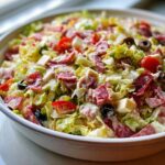 A close-up of a vibrant grinder salad tossed with creamy dressing, salami, cheese cubes, tomatoes, and olives in a white serving bowl.