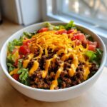 A close-up of a white bowl filled with a fresh taco salad, featuring seasoned ground beef, lettuce, tomatoes, shredded cheese, and cheese sauce.