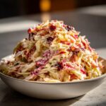 A large mound of creamy apple cranberry coleslaw featuring shredded cabbage and dried cranberries in a white bowl.