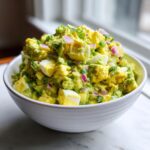 A white bowl filled with bright green avocado egg salad, featuring chunks of hard-boiled egg, red onion, and scallions.
