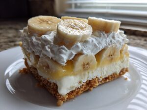 A square slice of rich banana cream cheesecake with a graham cracker crust, cream cheese filling, and topped with whipped cream and fresh banana slices.