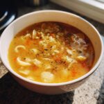 A close-up of a white bowl filled with golden pastina soup, featuring small pasta shapes and topped with dried herbs.