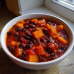 A close-up of a white bowl filled with rich, red butternut squash chili featuring large chunks of orange squash and black beans.
