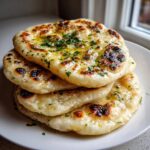 A stack of four freshly made, soft naan bread pieces brushed with melted butter and topped with chopped green herbs.