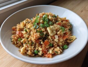 A close-up of a white bowl filled with savory cauliflower fried rice, featuring scrambled egg, carrots, peas, and green onions.