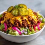 A close-up of a cheeseburger bowl featuring seasoned ground beef, melted cheese sauce, pickles, and red onion over lettuce.