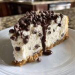 A decadent slice of chocolate chip cheesecake with a graham cracker crust, topped with mini chips and chocolate sauce.