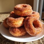 A stack of freshly made, golden brown mini donuts coated heavily in cinnamon sugar, resting on a white plate.