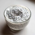 A glass filled with creamy coconut chia pudding, showing swollen chia seeds suspended in white liquid, topped with extra seeds.