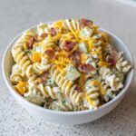 A close-up of a creamy pickle pasta salad made with rotini, topped with shredded cheddar cheese and crispy bacon bits.