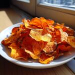 A white plate piled high with crispy, orange sweet potato chips seasoned with dark spices, sitting near a sunny window.