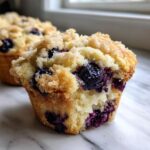 A close-up of a moist blueberry muffin featuring a sugary crumb topping and bursting with dark blueberries.