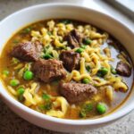 Close-up of a white bowl filled with rich broth, tender chunks of beef, egg noodles, and bright green peas in this easy beef noodle soup.