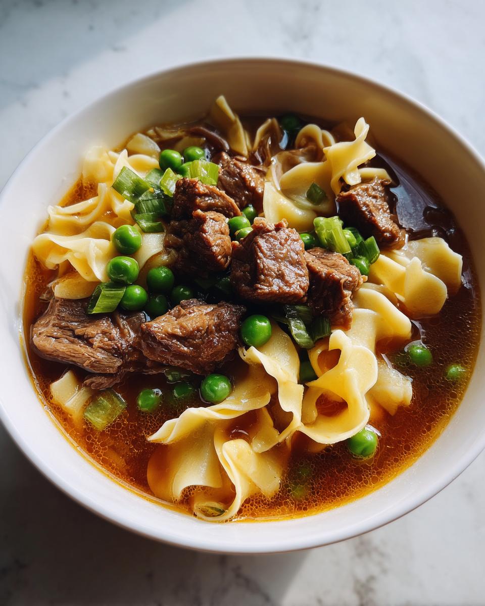 Close-up of a white bowl filled with rich beef noodle soup, featuring chunks of beef, egg noodles, peas, and green onions.