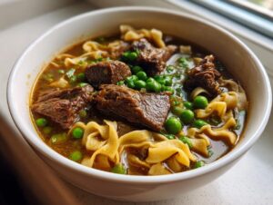 Close-up of a white bowl filled with rich beef noodle soup, featuring tender chunks of beef, wide egg noodles, and bright green peas.