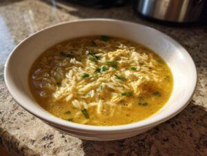 A close-up of a white bowl filled with golden pastina soup, featuring small pasta shapes and shredded chicken, garnished with parsley.