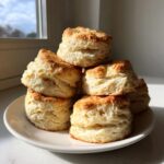 A stack of six golden-brown, flaky buttermilk biscuits piled high on a white plate near a window.