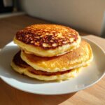 A stack of three golden-brown almond flour pancakes sitting on a white plate, beautifully lit by sunlight.