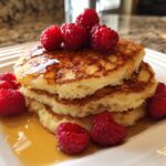 A stack of three fluffy ricotta pancakes drizzled with syrup and topped with fresh raspberries.
