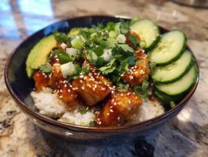 A close-up of a delicious salmon bowls featuring glazed salmon pieces over rice, topped with sesame seeds, cilantro, and served with sliced avocado and cucumber.