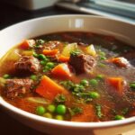 Close-up of a rich vegetable beef soup featuring tender beef chunks, bright orange carrots, green peas, and herbs.