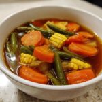 Close-up of a white bowl filled with rich, dark broth vegetable soup featuring large chunks of carrots, green beans, and corn on the cob pieces.