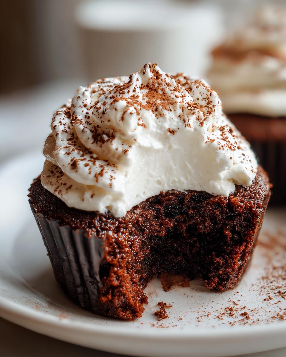 A close-up of a rich, dark chocolate hot chocolate cupcake with a bite taken out, topped with whipped cream and cocoa powder.
