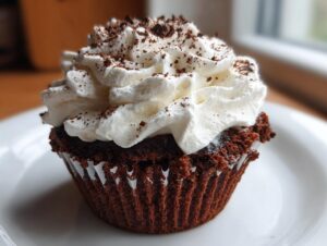 A close-up of a decadent hot chocolate cupcakes featuring a dark chocolate cake base topped with a swirl of white whipped cream and chocolate shavings.