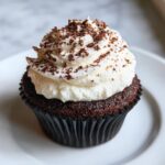Close-up of a rich, dark chocolate hot chocolate cupcake topped with thick white frosting and chocolate shavings.