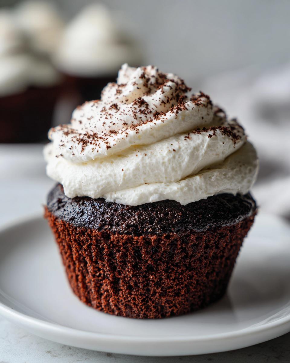 A close-up of a dark chocolate hot chocolate cupcake topped with a swirl of white whipped cream and chocolate shavings.