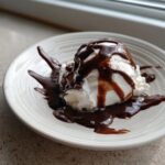 A scoop of vanilla ice cream generously covered in rich, dark chocolate sauce recipe, served on a white plate.