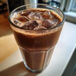 Close-up of a tall, sweating glass filled with iced chocolate milk and large ice cubes.