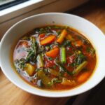 Close-up of a white bowl filled with rich, tomato-based minestrone soup featuring carrots, celery, and dark leafy greens.