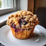A close-up of a moist blueberry muffin topped with a thick, sugary crumb topping, sitting on a white plate.