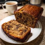 A freshly baked loaf of moist date nut bread, with one thick slice cut and resting on a white plate next to a mug.