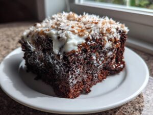 A moist, dark slice of german chocolate poke cake topped with white frosting and toasted coconut flakes.