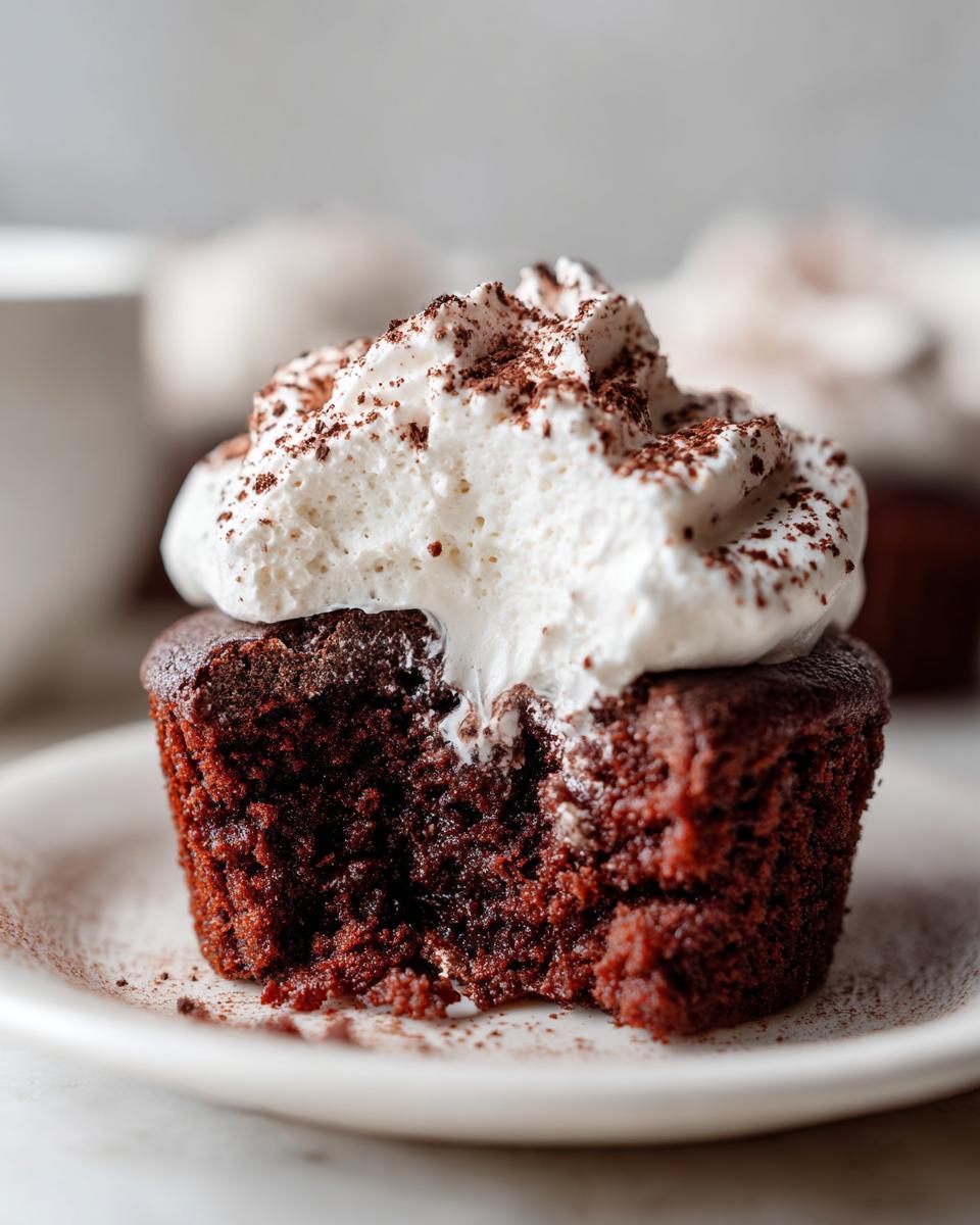 Close-up of a moist hot chocolate cupcakes with a bite taken out, topped with whipped cream and cocoa powder.