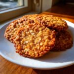 Close-up of several golden brown oatmeal lace cookies stacked on a white plate, catching bright sunlight.