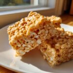 Two gooey squares of peanut butter rice krispie treats stacked slightly on a white plate, catching sunlight.