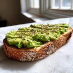 Close-up of a slice of toasted sourdough bread topped with mashed avocado and black pepper for perfect avocado toast.